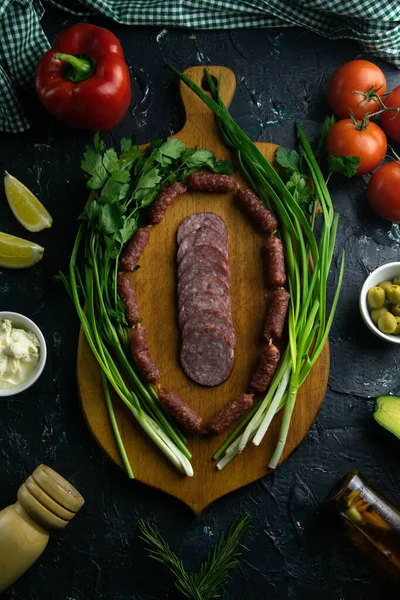 Salami rebanado en una tabla de madera, alrededor del jamón, cebollas verdes, eneldo y perejil . — Foto de Stock