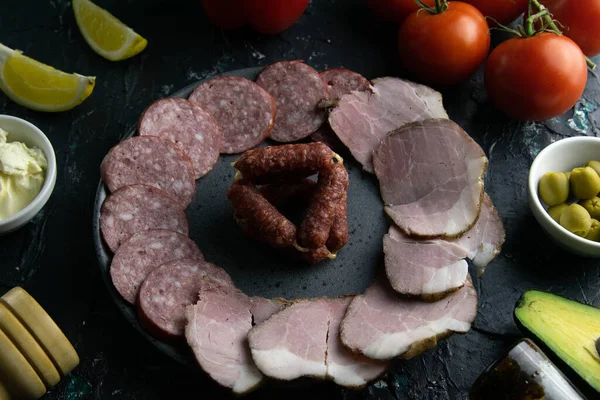 Plato gris con salchichas sobre fondo negro, alrededor del salami y jamón, tomate, souce, aceitunas, limón, aceite de oliva con especias . — Foto de Stock