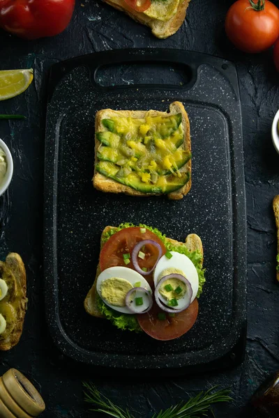 Grey cutting board with sandwich with tomato, egg and purple onion and avocado and cheese sandwich