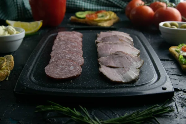 Tabla de cortar gris con salami y jamón. Alrededor de tomate, souce, aceitunas, limón, aceite de oliva con especias y sándwiches — Foto de Stock