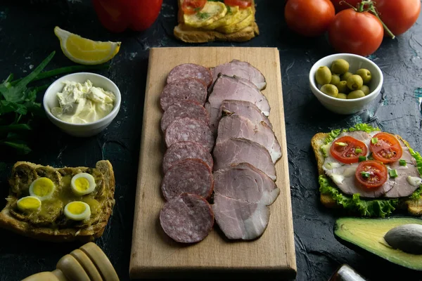 Salami en rodajas y jamón en una tabla de madera, alrededor de la tabla de aceitunas con otros sándwiches, tomate, limón y salsa . — Foto de Stock