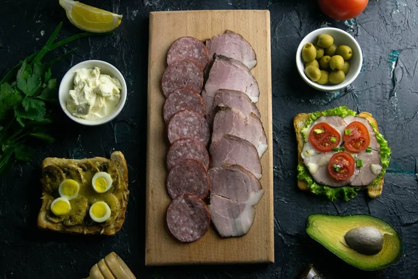 Salami en rodajas y jamón en una tabla de madera, alrededor de la tabla de aceitunas con otros sándwiches, tomate, limón y salsa . — Foto de Stock