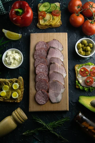 Salami en rodajas y jamón en una tabla de madera, alrededor de la tabla de aceitunas con otros sándwiches, tomate, limón y salsa . — Foto de Stock