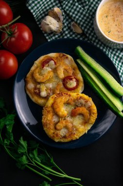 Mavi tabakta karides ve peynirli küçük bir pizza. Bir tabak domates, sos, otlar ve çatal bıçak takımı etrafında salatalık.