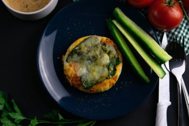 Mavi tabakta ıspanaklı ve peynirli küçük bir pizza. Bir tabak domates, sos, otlar ve çatal bıçak takımı etrafında salatalık.