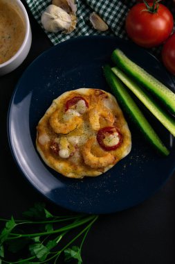 Mavi tabakta karides, domates ve peynirli küçük bir pizza. Bir tabak domates, sos, otlar ve çatal bıçak takımı etrafında salatalık.