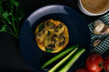 Mantarlı küçük bir pizza ve mavi tabakta peynirli salatalık, sos, şifalı otlar ve çatal bıçak takımı.