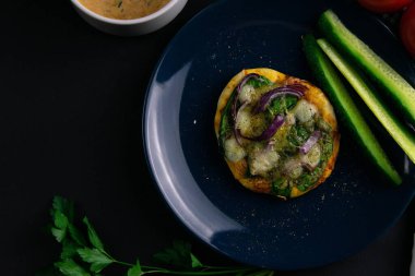 Mavi tabakta ıspanaklı, mor soğanlı ve peynirli küçük bir pizza. Bir tabak domates, sos, otlar ve çatal bıçak takımı etrafında salatalık.