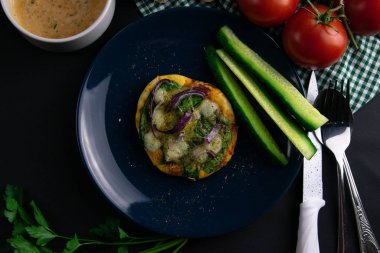 Mavi tabakta ıspanaklı, mor soğanlı ve peynirli küçük bir pizza. Bir tabak domates, sos, otlar ve çatal bıçak takımı etrafında salatalık.