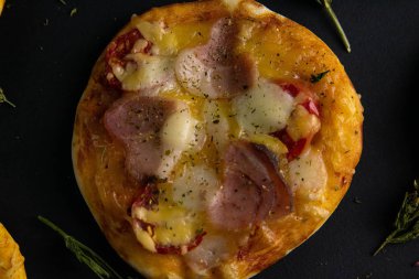Küçük mini pizza. Siyah arka planda jambonlu ve peynirli pizza. Yakınlarda başka pizzalar da görünüyor..