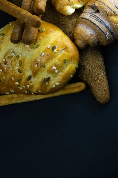 Különböző típusú friss kenyér gabonával fekete rusztikus háttérrel. — Stock Fotó
