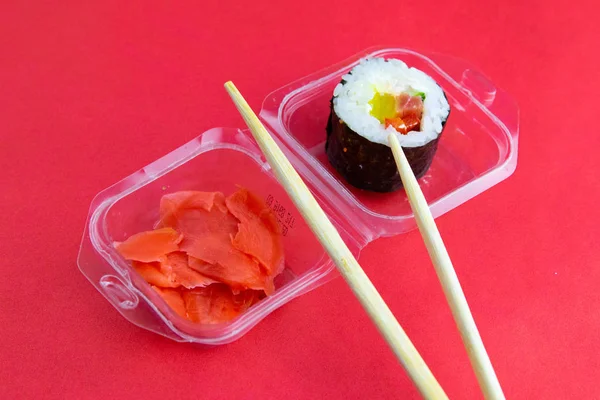 Sticks to reach for the roll, roll in the piece of packaging, next to the role of pickled ginger, everything stands on a red background