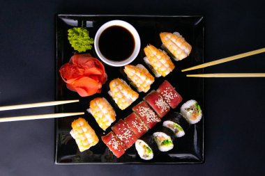 Suşi, soya sosu, kırmızı marinaginger ve soya sosu için çubukların yanında duran farklı rulolarla dolu siyah tabak..