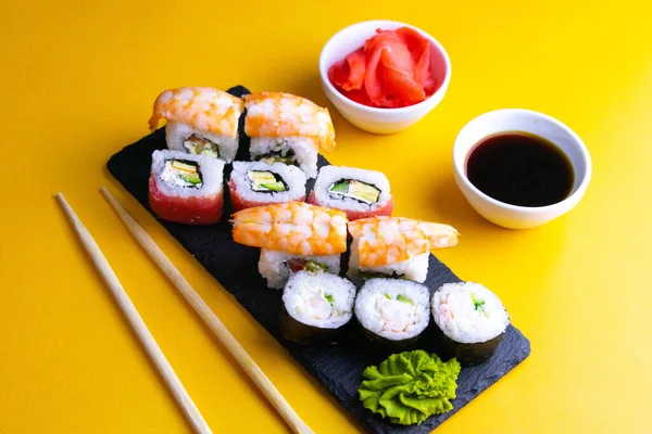 Granite board for sushi, filled with rolls of different kinds, next to stand sticks for sushi, red marinaginger and soy sauce. — Stock Photo, Image