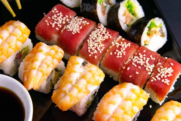 Czarna płyta z sushi, wypełniona rolkami różnego rodzaju, obok stojaków na sushi, czerwony marinaginger i sos sojowy. — Zdjęcie stockowe