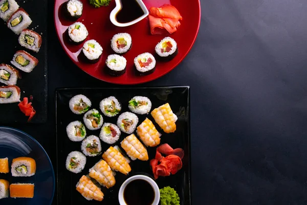 Különböző típusú tekercseket helyeznek el különböző tányérokon, szójaszósszal, wasabival és sushi pálcikákkal együtt, a tányérok fekete alapon állnak. — Stock Fotó