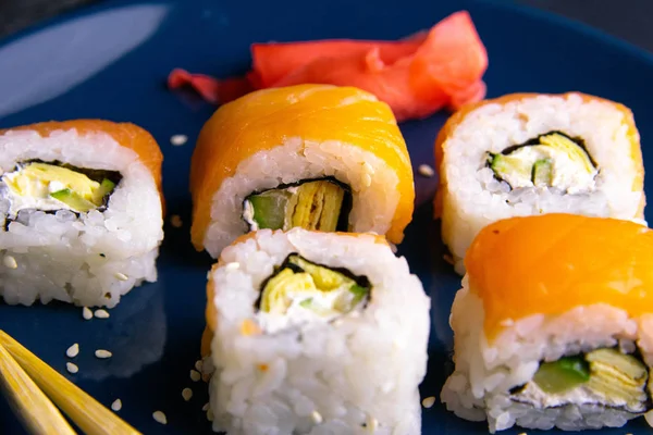Rolls na niebieskim talerzu wraz z marynowanym czerwonym imbirem, obok znajduje się sos sojowy i kije do sushi — Zdjęcie stockowe