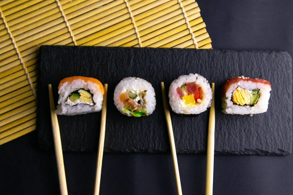 Kije trzymać rolki, które stoją na czarnej tablicy grafitowej do sushi, który stoi na czarnym tle i rolki na sushi spin — Zdjęcie stockowe