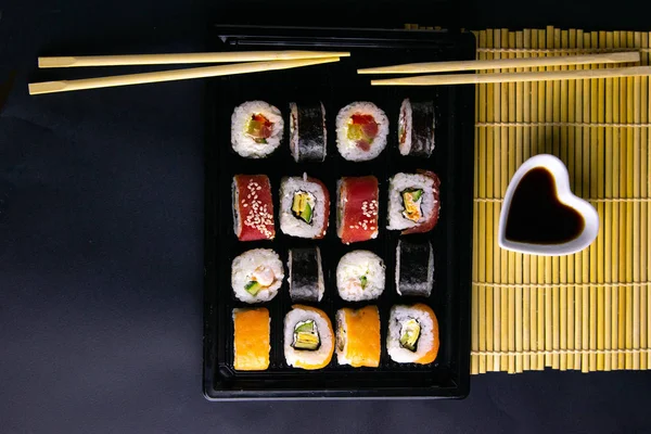 Różne rodzaje rolki stoją na czarnej płycie, która stoi na czarnym tle, obok dywanu do spinning rolki stoi sos sojowy i kije do sushi — Zdjęcie stockowe