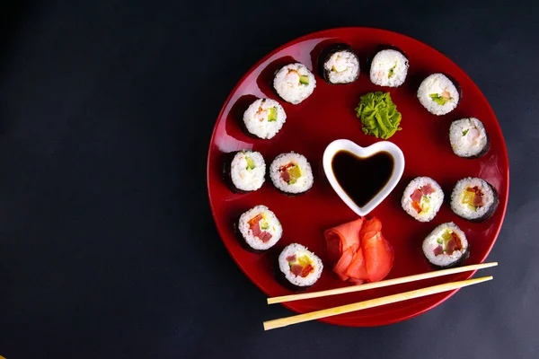 Rolki różnego rodzaju są układane na czerwonej płycie wraz z sosem sojowym, wasabi i sushi patyczki, płyta stoi na czarnym tle — Zdjęcie stockowe