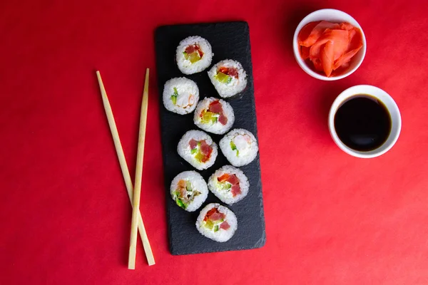 寿司のための花崗岩板に赤とオレンジのロール,寿司のための棒の横に,生姜と醤油,すべての赤の背景に立って. ロイヤリティフリーのストック画像