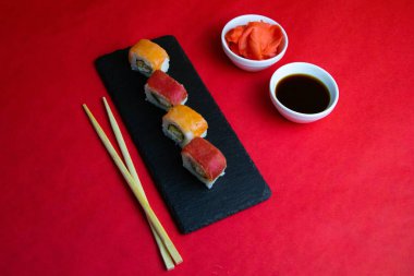 Suşi, zencefil ve soya sosu çubuklarının yanında, kırmızı arka planda duran granit bir tahtanın üzerinde kırmızı ve turuncu rulo.