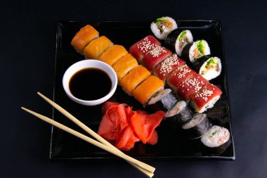 Suşi, soya sosu, kırmızı marinaginger ve soya sosu için çubukların yanında duran farklı rulolarla dolu siyah tabak..