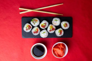 Suşi, zencefil ve soya sosu çubuklarının yanında, kırmızı arka planda duran granit bir tahtanın üzerinde kırmızı ve turuncu rulo.