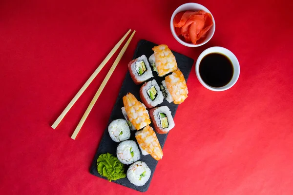 Granite board for sushi, filled with rolls of different kinds, next to stand sticks for sushi, red marinaginger and soy sauce. — Stock Photo, Image