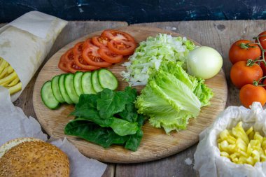 Hamburger hazırlıklarına hazırlanıyorum. Tahtada marul yaprakları var, doğranmış: domates, salatalık, soğan, pirzola ve sos..