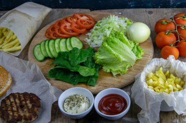 Hamburger hazırlıklarına hazırlanıyorum. Tahtada marul yaprakları var, doğranmış: domates, salatalık, soğan, pirzola ve sos..