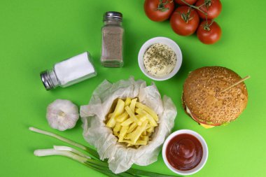 Yeşil arka planda hamburgerler, yakınlarda patates kızartmaları, soslar ve domateslerin yanı sıra tuzlu ve biberli mısırlar var..