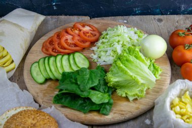Hamburger hazırlıklarına hazırlanıyorum. Tahtada marul yaprakları var, doğranmış: domates, salatalık, soğan, pirzola ve sos..