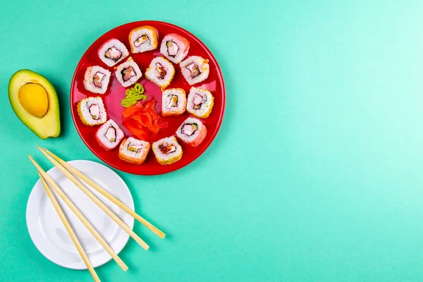 Red plate with sushi, filled with rolls of different kinds, next to stand sticks for sushi, red marinaginger and soy sauce on a mint background. Top views