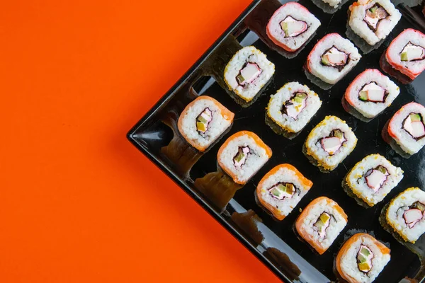 Black plate with sushi, filled with rolls of different kinds, next to stand sticks for sushi, red marinaginger and soy sauce on a orange background. Top views with clear space