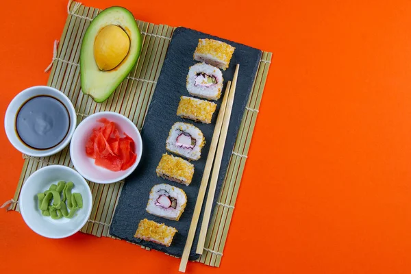 Granit board with sushi, filled with rolls of different kinds, next to stand sticks for sushi, red marinaginger and soy sauce on orange bacground. Top views with clear space