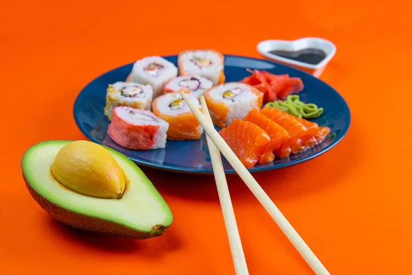 Blue plate with sushi, filled with rolls of different kinds, next to stand sticks for sushi, red marinaginger and soy sauce. on a orange background. Front views