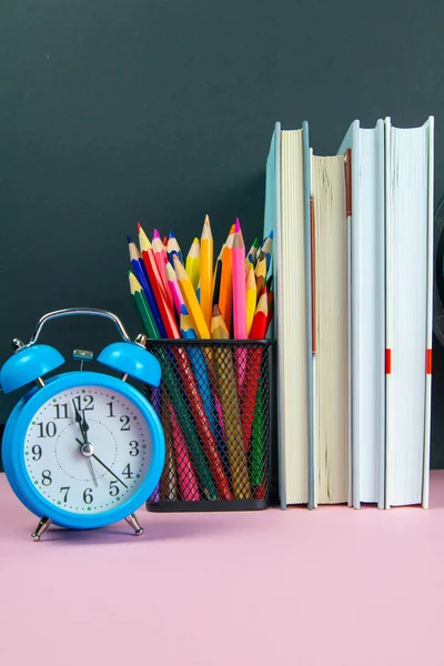 A stack of books, a clock and a glass with multi-colored pencils.