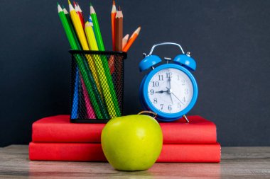 Mavi çalar saat, çok renkli kalemlerle birlikte kırmızı kitapların üzerinde duruyor, bir elmanın yanında..