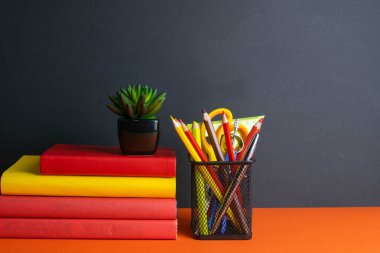 Bir yığın kitap, yeşil bir çiçek ve turuncu arka planda duran bir bardak kalem..