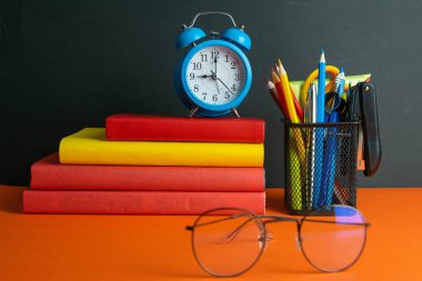 Bir yığın kitap, saat, dünya küresi ve ön planda kadın gözlükleri turuncu bir arka planda duruyor..