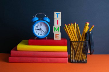 Bir yığın kitap, bir yeşil çiçek, mavi saat ve bir bardak kalem, ve turuncu bir arka planda yazı saati olan tahta küpler..