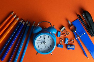 Kalemler, mavi alarm saati, pusula, cetvel ve zımba turuncu arka planda..