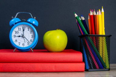 Mavi çalar saat, çok renkli kalemlerle birlikte kırmızı kitapların üzerinde duruyor, bir elmanın yanında..