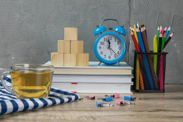 A stack of books, a clock, wooden cubes in the shape of a pyramid, next to there is tea with lemon in a transparent glass glass and multi-colored pencils.
