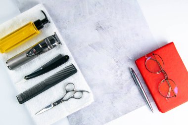 Saç kesimi, makas, tarak, tehlikeli jilet ve jöle makinesi beyaz havlunun üzerinde duruyor ve yanında da kadın gözlükleri olan bir kalem ve kırmızı defter var..