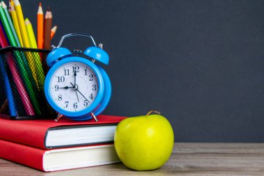 Mavi çalar saat, çok renkli kalemlerle birlikte kırmızı kitapların üzerinde duruyor, bir elmanın yanında..