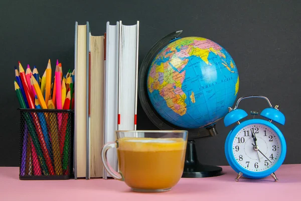 Coffe is in the foreground and behind him are a stack of books, a globe, a clock and a glass of pencils.