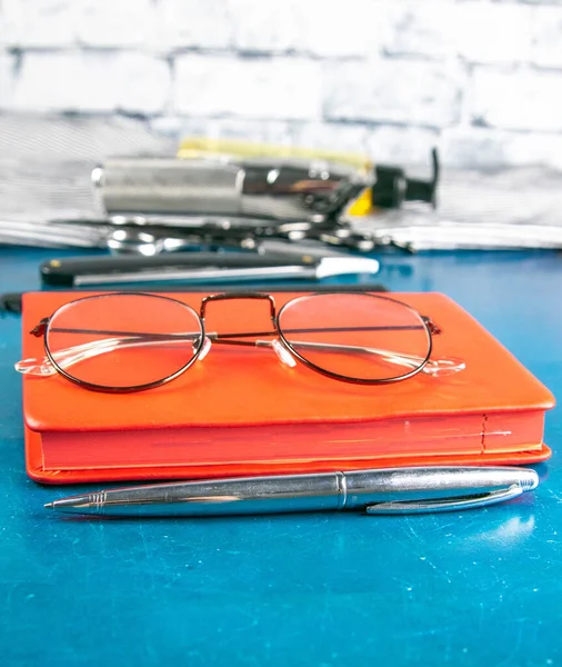 Hairdressing tools set, Red notepad with glasses and scissors, hair clipper, comb, dangerous razor, hair gel stand on blue background.