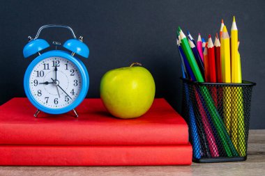 Mavi çalar saat, çok renkli kalemlerle birlikte kırmızı kitapların üzerinde duruyor, bir elmanın yanında..